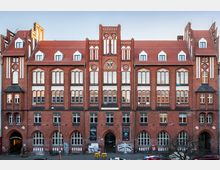 Backstein-Gebäude im neugotischen Stil mit symmetrischer Fassade, spitzbogigen Fenstern und einem roten Ziegeldach. Das Gebäude hat mehrere Giebel und ein zentrales Wappen oberhalb des Haupteingangs, der von weiteren Arkadenbögen flankiert ist.