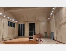 Innenraum einer großen, hellen Halle mit hoher Decke und cremefarbenen Wänden. Der Raum zeigt eine erhöhte Holzplattform mit Geländer, hohe Fenster mit grünen Rahmen, sowie einige gelagerte Gegenstände wie Kartons und Geräte; der Boden ist mit einem geometrischen Fliesenmuster gestaltet.