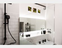 Modernes Badezimmer mit weiß gefliesten Wänden und einem großen beleuchteten Wandspiegel. Sichtbar sind ein schwarzes Duschset, ein Waschbecken mit schwarzem Wasserhahn, dekorative Pflanzen sowie Regale mit Handtüchern im Hintergrund.