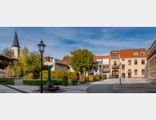 Kleiner, gepflasterter Platz in einer Altstadt mit umliegenden historischen Gebäude, hellen Fassaden und rotem Ziegeldach. Im Hintergrund ist ein Kirchturm sichtbar, daneben Bäume und gepflegte Grünanlagen.