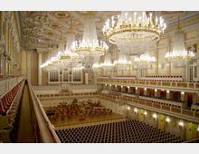 Prächtiger Konzertsaal mit reich verzierten goldenen und weißen Wänden, großen Kronleuchtern und einer Bühne voller Stühle und Musikpulte. Umlaufende Balkone mit roten Polstersitzen bieten Sitzplätze, und eine große Orgel ist in der Rückwand integriert.