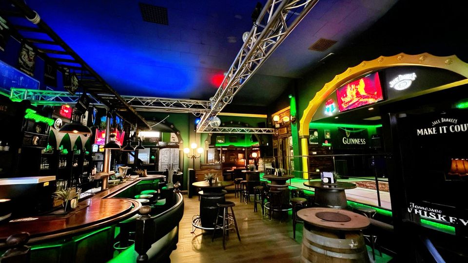 Innenansicht einer Bar im Pub-Stil mit geschwungener Theke, hohen Barhockern und mehreren Stehtischen auf Holzboden. Grüne und blaue Akzentbeleuchtung, Metall-Traversen an der Decke sowie Werbeschilder und Dekorelemente prägen den Raum.