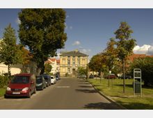 Straßenszene in einem Wohngebiet mit einem gelblichen historischen Gebäude im Hintergrund. Links parkende Autos entlang der Straße, rechts eine Rasenfläche mit jungen Bäumen und einem Mülleimer im Vordergrund.