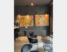 Ein modernes Café mit dunklen Wänden und minimalistischer Einrichtung. An der Wand hängen zwei bunte, abstrakte Gemälde, darunter ein Heizkörper; der Raum enthält schwarze Samtstühle und Metall-Beistelltische. Deckenlampen mit warmem Licht betonen die Kunstwerke.