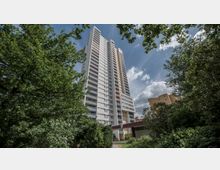 Hochhaus mit weißer Fassade und Balkonen, umgeben von dichtem Grün und Bäumen, unter blauem Himmel mit vereinzelten Wolken; im Vordergrund ein Eingangsbereich und gepflegte Vegetation.