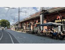 Straßenansicht mit einem historischen Backsteinviadukt, unter dem ein Irish Pub mit Außengastronomie eingerichtet ist. Vor dem Gebäude stehen Tische und Stühle, während im Vordergrund Straßenbahnschienen und ein gepflasterter Weg zu sehen sind.