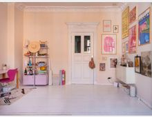 Helles Wohnzimmer mit pastellfarbenen Wänden, weiß gestrichenen Holzdielen und einer verzierten Stuckdecke. Der Raum enthält eine weiße Tür mit Spiegelglas, ein Regal mit Büchern und Dekorationen, eine pinkfarbene Sitzgelegenheit, und eine Wand mit gerahmten Kunstwerken und Postern.