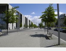 Breiter, gepflasterter Gehweg mit Reihen junger Bäume und Sitzbänken, flankiert von moderner Architektur aus Glas und Beton auf der linken Seite und einem historischen Gebäude mit Glaskuppel rechts; im Hintergrund ist der Berliner Fernsehturm sichtbar.