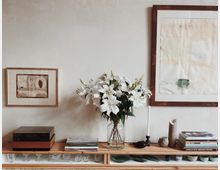 Ein minimalistisch eingerichtetes Regal vor einer cremefarbenen Wand, dekoriert mit weißen Lilien in einer Glasvase, Büchern und einer schwarzen Kerze. An der Wand hängen zwei gerahmte Kunstwerke, eines auf jeder Seite des Blumenarrangements.