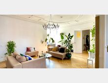 Wohnzimmer mit hohen Decken, einer weißen Wand, verzierten Stuckelementen und einem schwarzen Kronleuchter. Der Raum verfügt über helle Holzböden, beigefarbene Sofas, viele grüne Zimmerpflanzen und große Fenster, die Tageslicht hereinlassen.