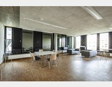 Moderner Konferenz- oder Aufenthaltsraum mit großen Fenstern, die viel Tageslicht hereinlassen. Der Raum verfügt über Holzfußboden, eine Tischgruppe mit Stühlen sowie eine Sitzecke mit grauen Sofas; die Decke besteht aus Akustikplatten.