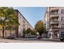 Straßenkreuzung in einem Wohngebiet mit mehrstöckigen Gebäuden im Stil der Nachkriegsarchitektur auf der linken Seite und Altbauten mit Balkonen auf der rechten Seite. Parkende Autos säumen die Straße, Bäume mit Herbstlaub stehen entlang der Gehwege, und ein Geschäft mit Schaufenster und ein Café befinden sich in den Erdgeschossen der Gebäude.