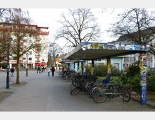 Straßenszene mit einem offenen Fahrradunterstand, dessen Stützen und Dach mit Plakaten und Graffiti bedeckt sind, daneben eine Reihe geparkter Fahrräder. Im Hintergrund ist ein Platz mit Bäumen, Menschen und einem Wohngebäude mit roten Balkonakzenten zu sehen.