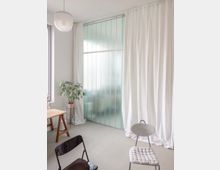 Heller, minimalistisch eingerichteter Innenraum mit großem Fenster bzw. milchiger Glasfront und bodenlangen weißen Vorhängen. Links stehen ein Holztisch und eine Topfpflanze, im Vordergrund sind zwei Stühle auf einem hellen Boden zu sehen.