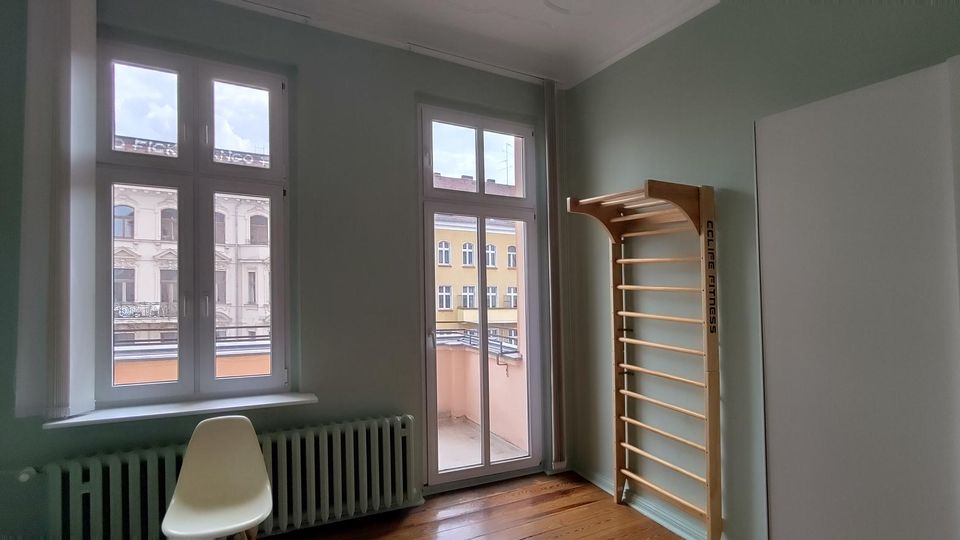 Heller Innenraum mit hohen Fenstern und einer Glastür zum kleinen Balkon, darunter ein Heizkörper und ein Stuhl. Rechts stehen eine Sprossenwand aus Holz und ein großer weißer Schrank vor grün gestrichenen Wänden und Holzboden.