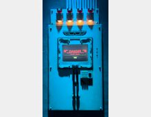 Ein technisches Bedienfeld an einer Wand, beleuchtet von blauem Licht, mit roten Anzeigen und einem Bildschirm mit der Warnung „DANGER - Emergency Destruction System“. Oberhalb befindet sich das Logo von „Arumo Industries“, darunter liegen Schläuche und eine Tastatur.