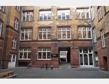 Backsteingebäude mit Innenhof, geprägt von gelb-rotem Klinker und großen weißen Fenstern. Im Erdgeschoss befindet sich ein Durchgang, daneben Türen, davor stehen mehrere Fahrräder auf gepflastertem Boden.