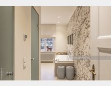 Helles Badezimmer mit einer freistehenden Badewanne, gemusterter Tapete an einer Wand und einer weißen Heizung unter einem Fenster mit hellen Vorhängen. Zwei dekorative Hocker stehen vor der Badewanne, und auf der Fensterbank befindet sich eine kleine Holzfigur.