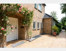 Backsteinfassade eines Hauses mit einem weißen Eingang, umgeben von rosafarbenen Blumenranken. Neben dem Eingang liegen mehrere schmale Fenster mit Gitterstäben, und der gepflasterte Weg führt an einem großen Pflanzkübel und Gartenhecken entlang.