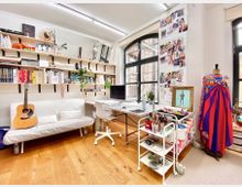 Helle und kreative Innenaufnahme eines Ateliers mit Holzboden. Zu sehen sind ein Sofa mit einer Akustikgitarre, ein Schreibtisch mit Computer, Regale mit Büchern und Dekor sowie ein Schneiderpuppe mit einem bunten Kleid. Ein Rollwagen mit Malutensilien steht neben weiteren Werkzeugen und einer Pinnwand voller Inspirationen.