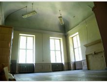 Ein leerer Raum mit hohen Decken, großen Fensterfronten mit Sprossen, verblassten Wänden und sichtbaren Rissen. Im Raum befinden sich Heizkörper unter den Fenstern, eine alte Kaminöffnung und abgehängte Deckenlampen.