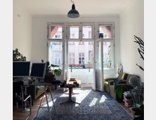 Wohnzimmer mit großen Fenstern, die auf einen kleinen Balkon führen. Der Raum ist mit einem Schreibtisch, zwei Monitoren, einem Teppich und einer grünen Couch eingerichtet und enthält mehrere Zimmerpflanzen.