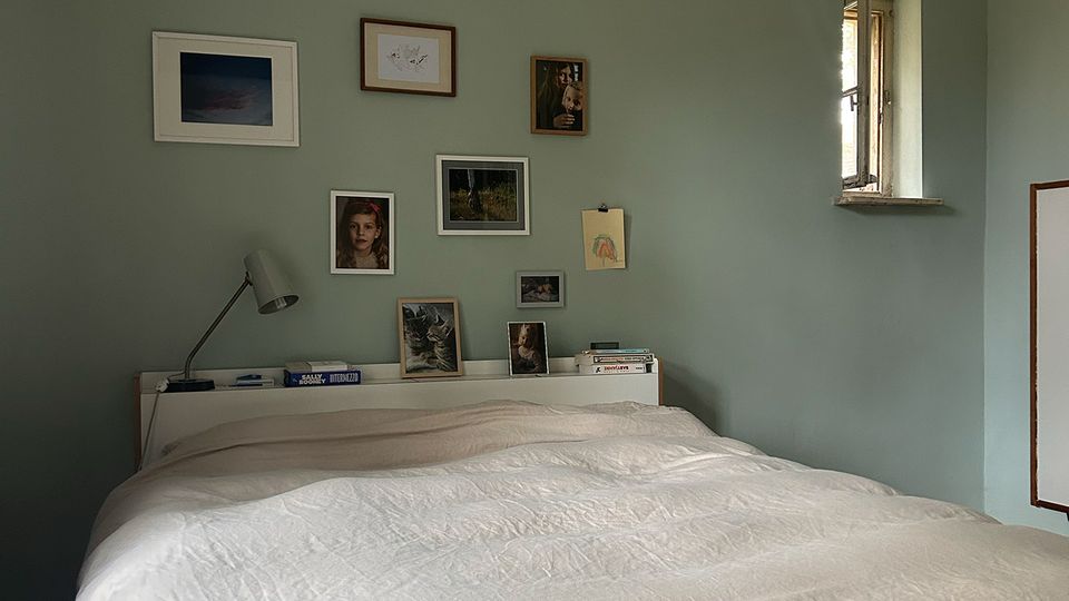 Schlafzimmer mit großem Bett und weißer Bettdecke vor einer salbeigrünen Wand, die mit mehreren gerahmten Bildern dekoriert ist. Neben dem Bett stehen eine schlichte Lampe und Bücher, rechts ist ein kleines tiefes Fenster in der Wand zu sehen.