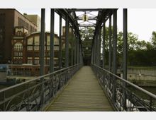 Eine schmale Fußgängerbrücke aus Metall mit Holzplanken erstreckt sich über einen Kanal. Im Hintergrund sind im Wasser liegende Gebäude im Backsteinstil mit verzierten Fassaden und große Bäume sichtbar.