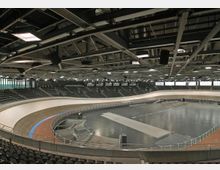 Innenansicht einer modernen Radrennbahn in einer großen Sporthalle mit geneigter Holzbahn, umgeben von Tribünen mit zahlreichen Sitzplätzen. Die Decke ist von einem komplexen Stahlträgernetz mit Beleuchtungselementen geprägt.