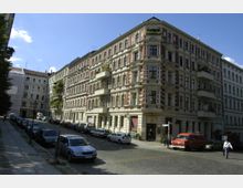 Straßenszene mit historischen Wohngebäuden im Gründerzeitstil, gekennzeichnet durch rot-weiße Fassaden, Balkone und ornamentale Verzierungen; parkende Autos säumen die gepflasterte Straße, während der Himmel klar und blau ist.