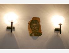 Eine Holz-Ikone mit dem Porträt eines bärtigen Mannes hängt an einer beige verputzten Wand, flankiert von zwei modernen Wandlampen mit runden, leuchtenden Glaskugeln.