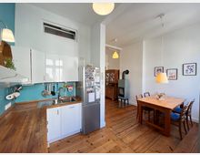 Wohnküche mit Holzfußboden, bestehend aus einer weißen Einbauküche mit türkisfarbener Wand, einem Esstisch mit Holzstühlen und Blumenarrangement sowie einem Kaminofen im Hintergrund. Die Wände sind weiß, dekoriert mit Bildern, und von der Decke hängen schlichte Lampen.