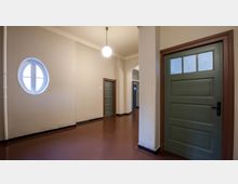 Ein einfacher Innenflur mit cremefarbenen Wänden und einem dunkelroten Boden. Rechts und links befinden sich grüne Türen mit Holzrahmen, und an der linken Wand ist ein rundes Fenster sichtbar. Eine einzelne Deckenlampe beleuchtet den Bereich.