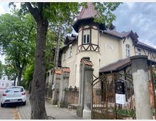 Straßenszene mit einem historischen Gebäude aus dem Jahr 1902, das im Jugendstil gestaltet ist, mit Fachwerkelementen, Erker und Satteldach. Ein schmiedeeisernes Tor und eine alte Steinmauer umgeben das Grundstück, während ein Baum und ein geparktes Auto auf dem Gehweg sichtbar sind.