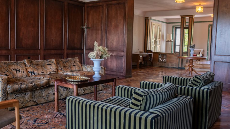 Wohnzimmer mit dunkler Holzvertäfelung an den Wänden, gepolsterten Möbeln mit gemusterten Stoffen, einem orientalischen Teppich auf Holzboden und einem kleinen Holztisch mit Blumenvase. Im Hintergrund ein angrenzender Raum mit großen Fenstern und klassischen Holzmöbeln.