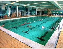 Hallenbad mit einem großen Schwimmbecken, in dem mehrere Personen schwimmen. Die Wände sind mit mosaikartigen Wellenmustern dekoriert, und am Beckenrand befinden sich Liegestühle und Sonnenschirme.