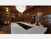 Holzvertäfelter Konferenzraum mit rechteckig angeordneten Tischen und Stühlen, darüber eine moderne, weiße Hängelampe. Im Hintergrund wandhohe Holzwände mit eingelassener Tür und einer Uhr.