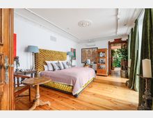 Schlafzimmer mit Holzboden, einem auffälligen gelb gemusterten Bettrahmen und einer rosafarbenen Tagesdecke. An den weißen Wänden befinden sich Kunstwerke, ein dekorativer Stuck ziert die Decke, und durch eine geöffnete Tür ist ein Raum mit viel Grünpflanzen sichtbar.