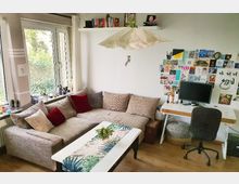 Wohnzimmer mit einer großen Eckcouch, dekoriert mit roten und beigen Kissen, einem weißen Couchtisch mit tropischem Muster und einer kleinen Grünpflanze. Rechts im Bild befinden sich ein Schreibtisch mit einem Computer und eine Pinnwand mit Karten und Fotos; im Hintergrund große Fenster mit Blick ins Grüne.