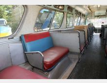 Innenraum eines alten Linienbusses mit rot-blauen Sitzen und Metallrahmen. Große Fenster an den Seiten lassen Licht herein, und der Boden besteht aus dunklem geriffelten Material.