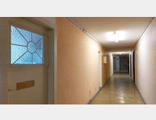 Ein langer, schmaler Flur mit hellorange gestrichenen Wänden, weißer Decke und abgenutztem Linoleumboden. Auf der linken Seite befindet sich eine Tür mit geriffeltem Glasfenster und einem Gitter, während der Flur durch Deckenleuchten beleuchtet wird.