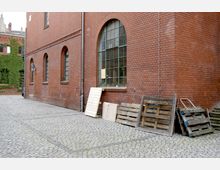 Backsteinfassade eines Industriegebäudes mit großen, halbrunden Fenstern auf einem gepflasterten Hof. An der Wand lehnen Holzpaletten und ein Metallgitter, im Hintergrund sind begrünte Wände zu sehen.