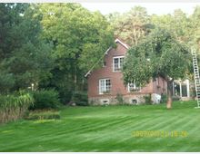 Backsteinhaus mit weißen Fenstergittern, umgeben von einem gepflegten Rasen, Bäumen und Sträuchern. Im Hintergrund ist dichter Wald zu sehen, und rechts steht eine Leiter an einem Baum.