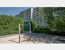 Spielplatz mit einer Schaukel auf einem Sandboden, umgeben von dichten grünen Hecken und Bäumen. Im Hintergrund steht ein mehrstöckiges Wohngebäude aus Beton mit gelben Akzenten.