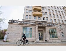 Fassade des Computerspielemuseums, einem Gebäude mit heller Steinverkleidung und großen Fenstern, verziert durch ein Vordach aus Metallstreben. Eine Person fährt in der Vordergrundszene auf einem Fahrrad vorbei.