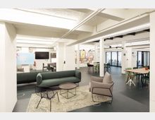 Großzügiger, moderner Gemeinschaftsraum mit Sitzgelegenheiten, darunter ein grünes Sofa, ein Sessel und ein runder Couchtisch auf einem Teppich. Im Hintergrund sind Tische mit Stühlen, ein wandmontierter Bildschirm und bunte Wanddekorationen zu sehen, umgeben von weißen Säulen und Glaswänden.