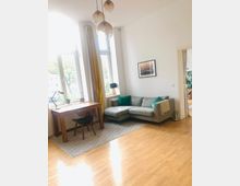 Helles Wohnzimmer mit Holzboden, einer grauen Eckcouch mit grünen Kissen und einem Schreibtisch mit Stuhl. Große Fenster mit gelben Vorhängen lassen viel Tageslicht herein, daneben hängt eine Pendelleuchte mit mehreren gläsernen Lampenschirmen.