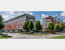 Wohnanlage mit mehrstöckigen, farblich gestalteten Gebäuden in Rot-, Weiß- und Grautönen, umgeben von Grünflächen und Bäumen. Im Vordergrund ist ein Gehweg, parkende Autos, sowie ein Verkehrsschild mit der Aufschrift „30 Zone“ zu sehen.