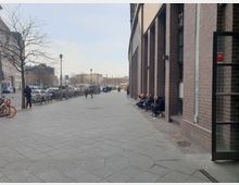 Eine städtische Straße mit breitem Gehweg und Fahrrädern an Ständern auf der linken Seite. Rechts ein langes Gebäude mit Backsteinfassade, vor dem mehrere Personen auf niedrigen Vorsprüngen sitzen, während im Hintergrund Autos und Menschen den offenen Platz überqueren.