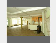 Großer, offener Raum mit hohen Decken, einem Sichtbeton-Pfeiler in der Mitte, Holzboden und einer Küchenzeile mit orangefarbener Wand im Hintergrund. Ein Kaminofen steht nahe einer Wand mit sichtbaren Ziegeln.